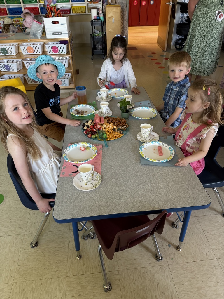 Kindergarten students and teachers dressed in spring hats enjoy a classroom tea party with snacks, tea cups, and colorful decorations.