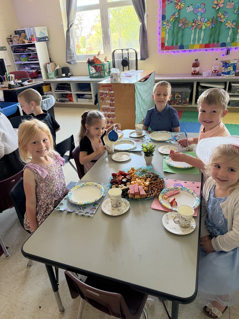 Kindergarten students and teachers dressed in spring hats enjoy a classroom tea party with snacks, tea cups, and colorful decorations.