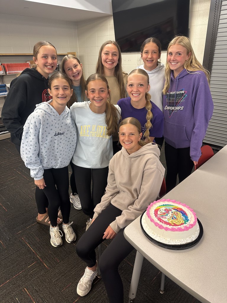 Nine middle school girls smile together in a classroom beside a Barbie-themed cake during a birthday celebration.