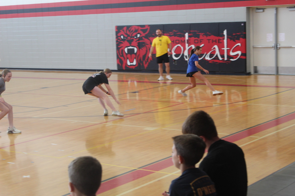 Students sprint and change direction during an agility drill in the school gym while classmates watch.
