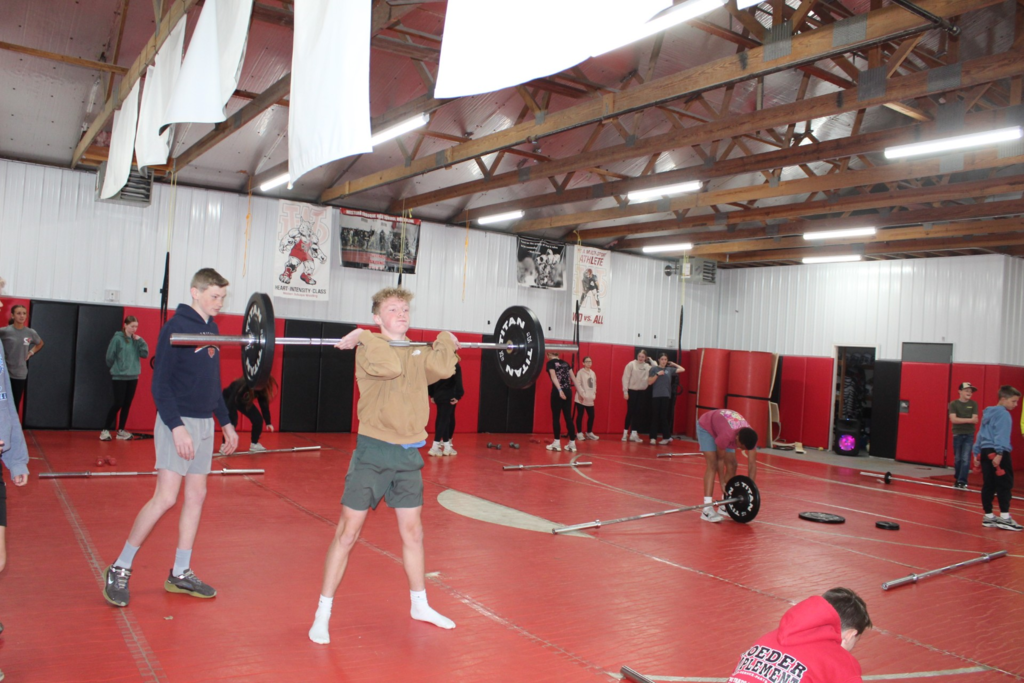 Student performs a barbell lift while classmates practice and observe during a physical fitness class.
