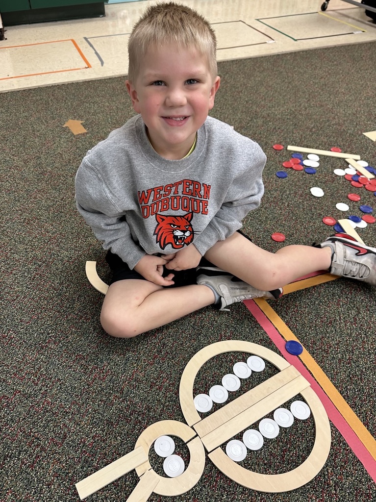 Young student smiles beside a large insect design created with wooden shapes and counting chips on the classroom floor.
