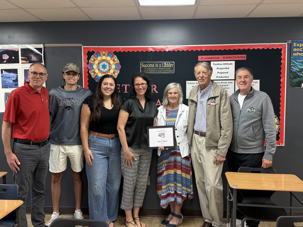 Seven adults and one student stand together in a classroom while presenting a recognition certificate during a school celebration.