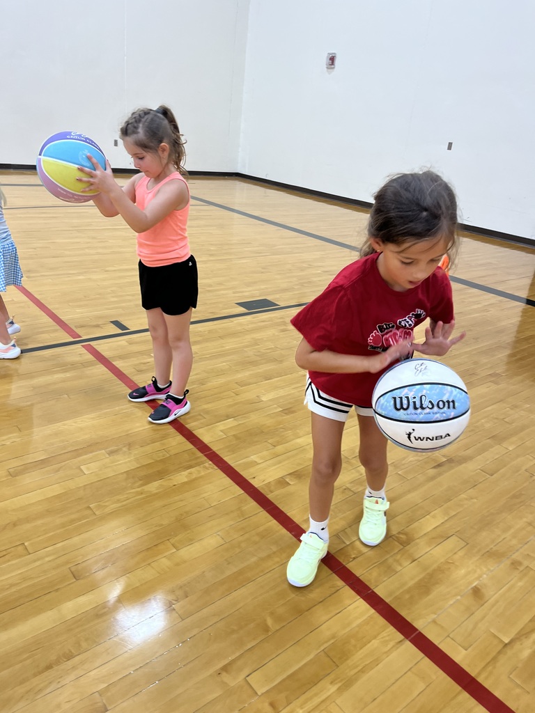 Two young girls practice basketball dribbling in a gym, focusing on ball control during a youth skills camp.