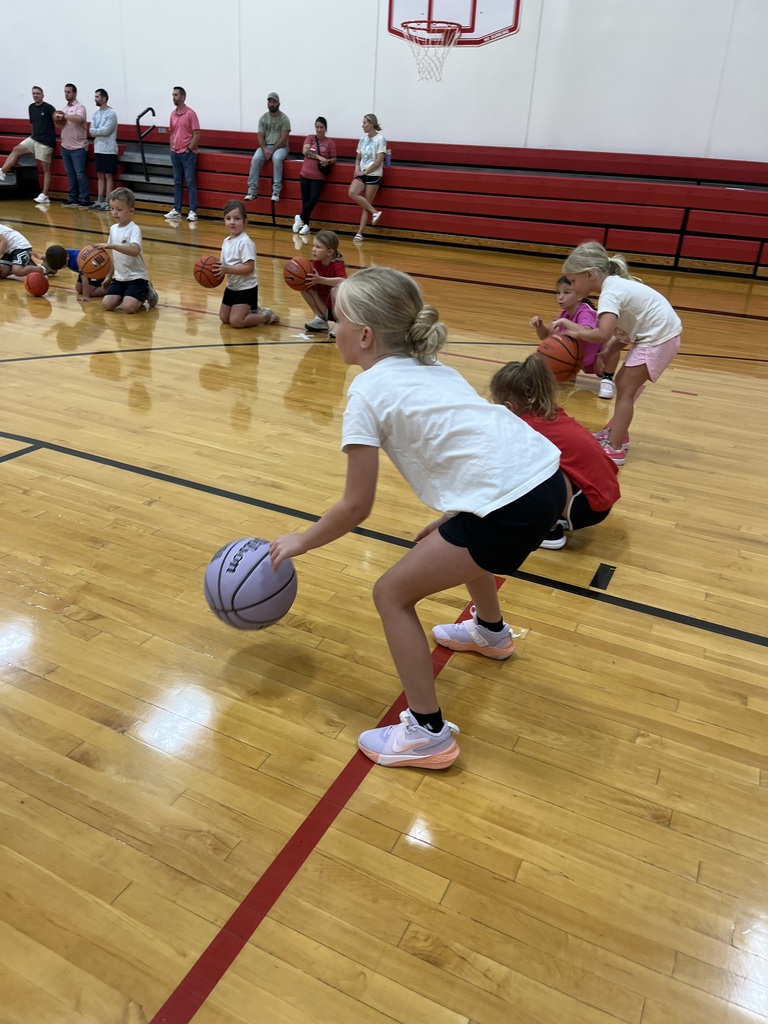 Young basketball campers kneel and dribble basketballs during a group skills drill while parents watch from the bleachers.
