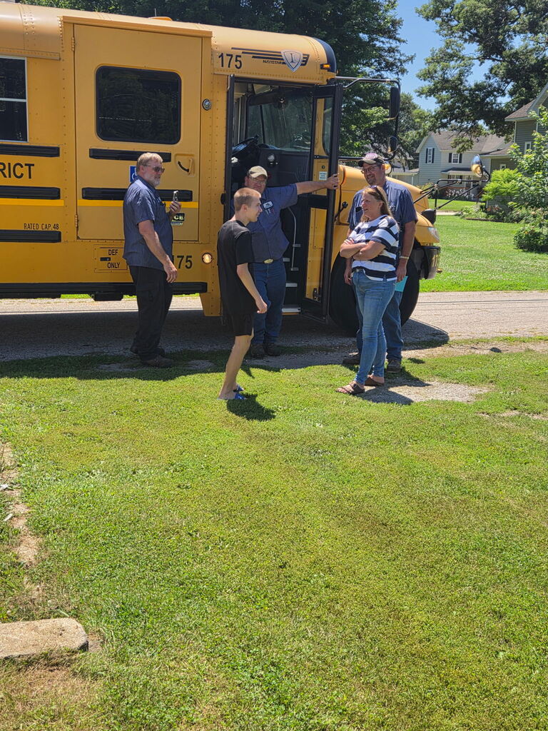 School transportation staff, a student, and family members gather beside a school bus and talk near the open bus door.