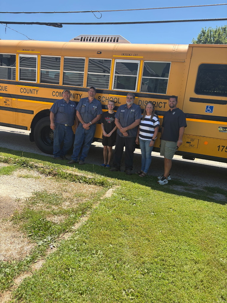 School transportation staff and a student stand beside a school bus during a community visit on a sunny day.