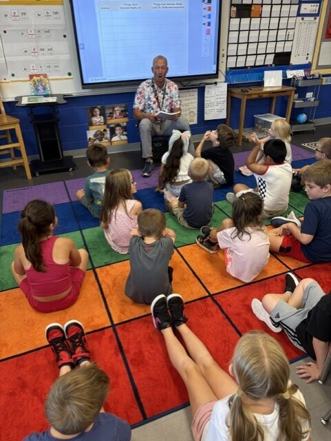 A teacher reads to elementary students seated together on a colorful classroom rug.
