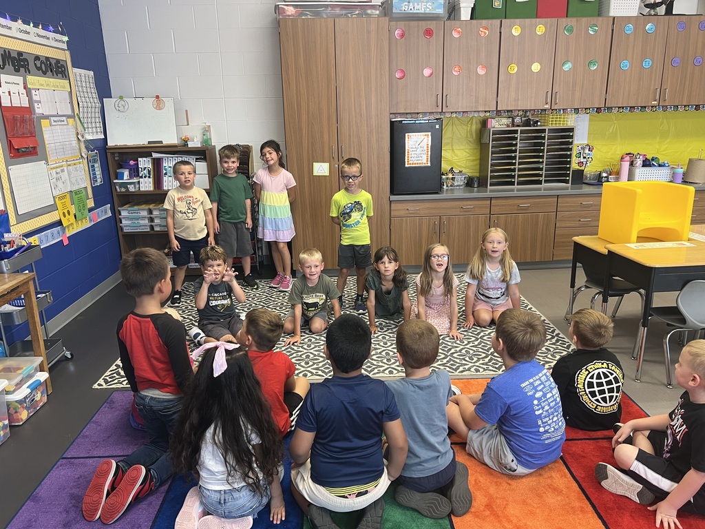 Elementary students gather on a colorful classroom rug, with several children standing in front of their seated classmates.