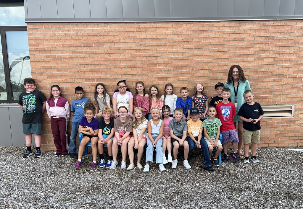A class of elementary students and their teacher pose together outside against a brick school wall.
