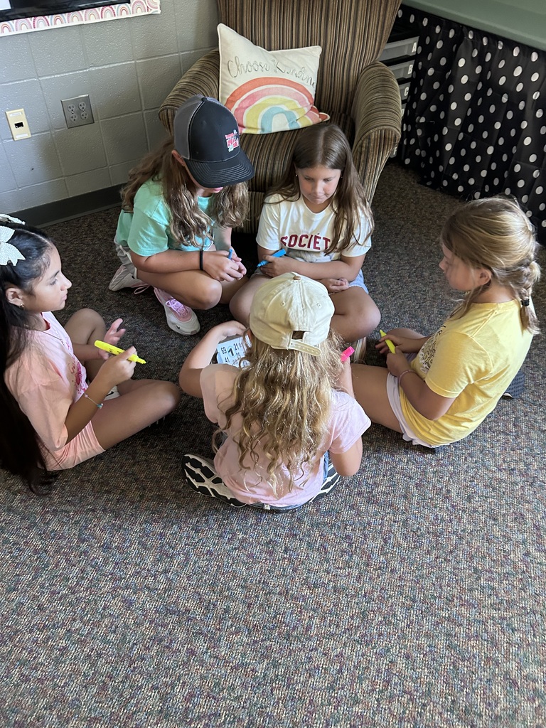 Five students sit in a circle on the classroom floor, using colorful markers during a small-group activity.
