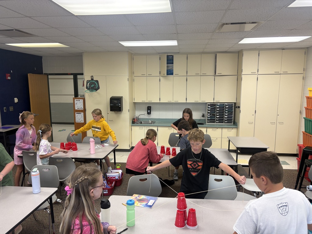 Students work in groups at classroom tables, using string and stacked red cups for a hands-on team-building activity.
