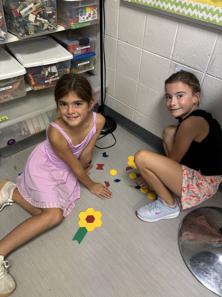 Two students sit on the classroom floor, smiling beside colorful pattern blocks arranged into a flower.