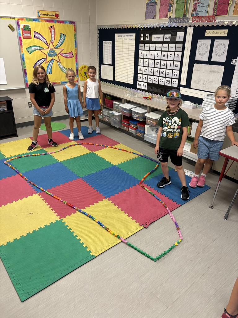 Five students stand beside a long, colorful chain they created across foam floor tiles in their classroom.