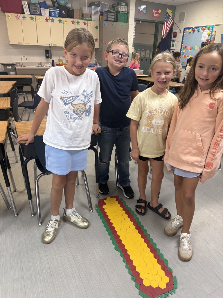Four students smile in their classroom beside a long design made from red, yellow, and green pattern blocks.