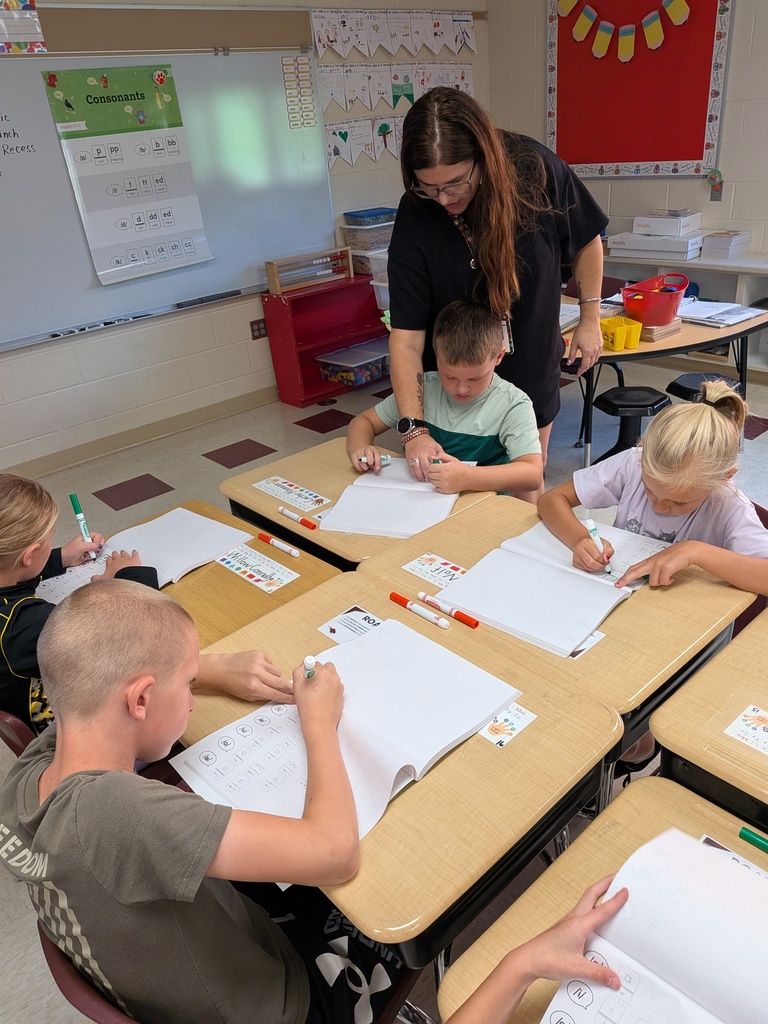 Students sitting at their desks writing with markers while a teacher helps them.