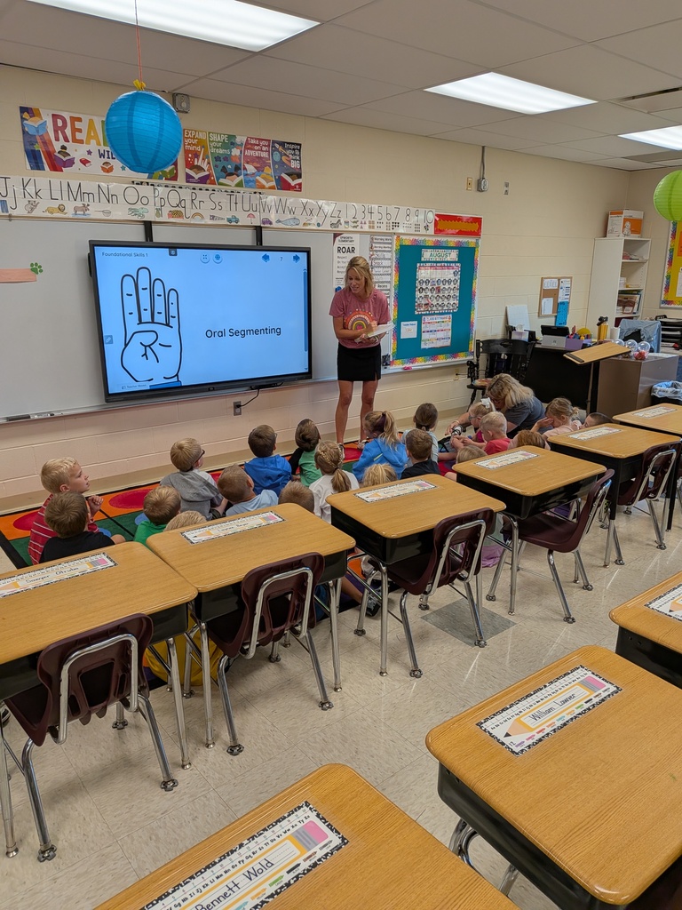 A classroom with students sitting on the floor listening to the teacher.