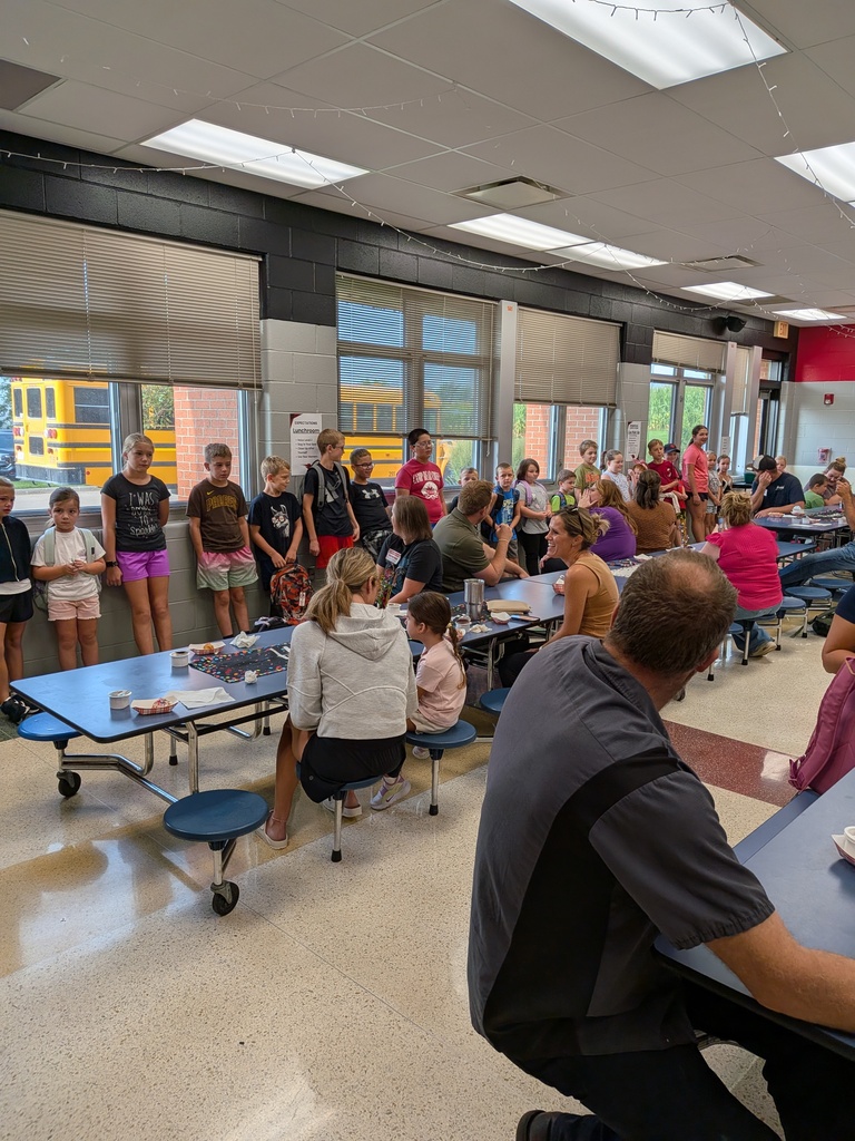 Students and adults gather around cafeteria tables during a school birthday celebration, with children lined along the wall.