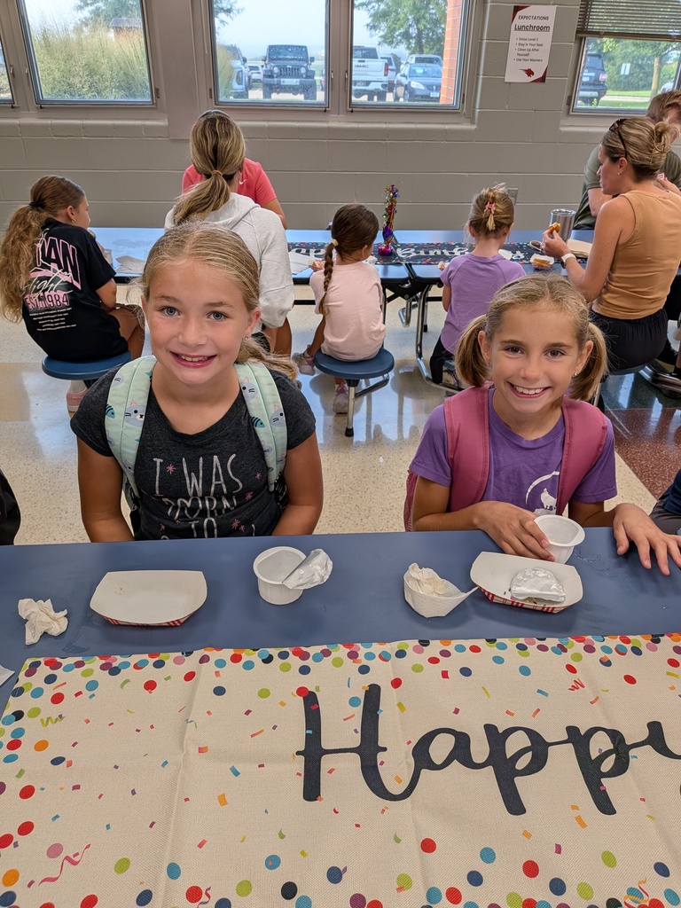 Two smiling students with backpacks sit at a cafeteria table decorated with a colorful “Happy” birthday table runner.