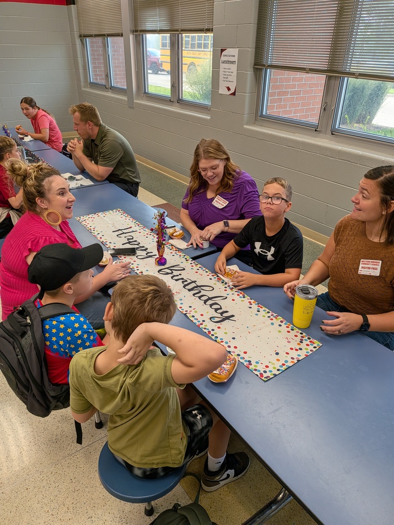 Students and school staff gather around a cafeteria table with treats and a colorful “Happy Birthday” runner.