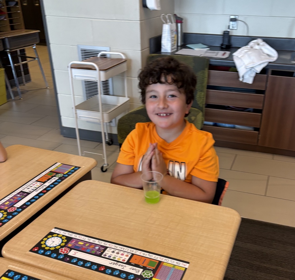 Smiling student in an orange shirt sits at a classroom desk with a small cup of bright green liquid.