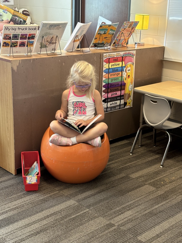 Student with glasses sits cross-legged on an orange ball chair, quietly reading a book in a classroom reading area.