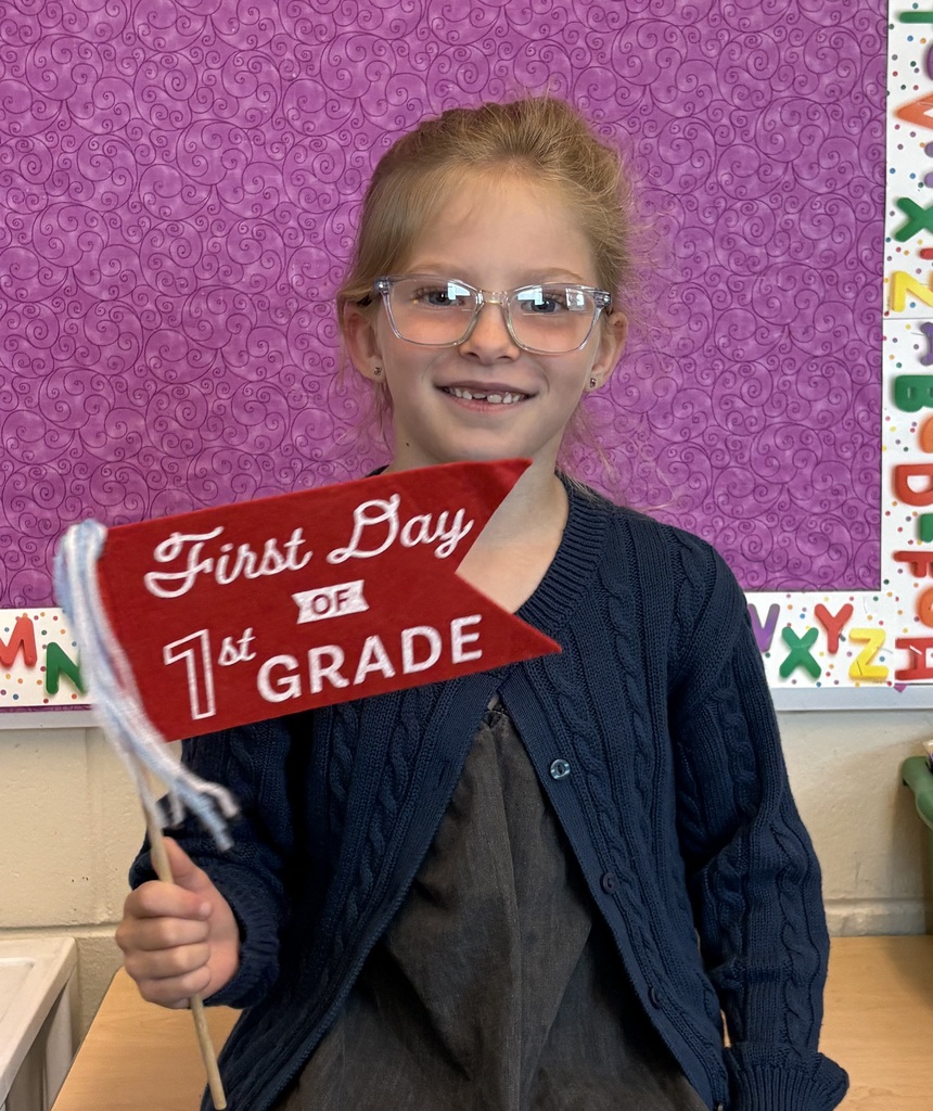 Smiling first-grade student with glasses holds a red pennant reading “First Day of 1st Grade” in a classroom.