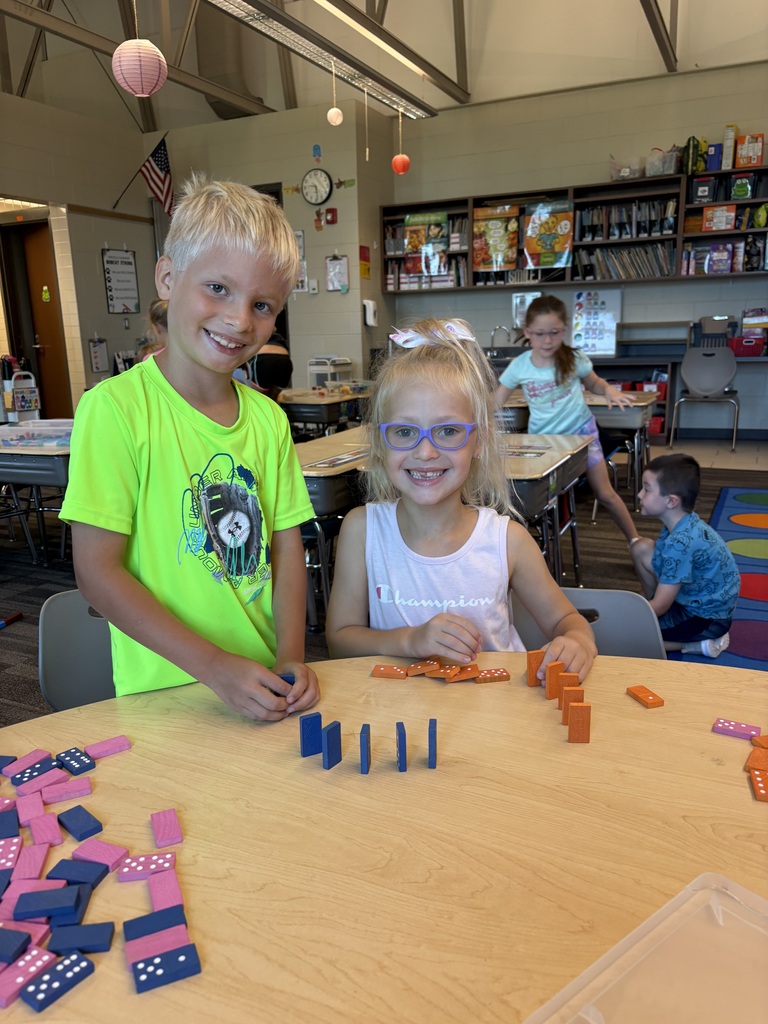 Two smiling students stand beside a classroom table, arranging colorful dominoes during a hands-on activity.
