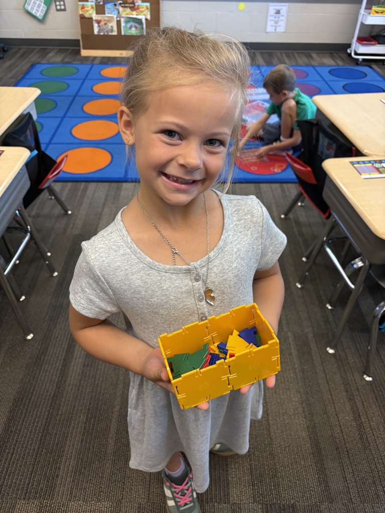 Smiling student holds a yellow container filled with colorful geometric pieces during a classroom activity.