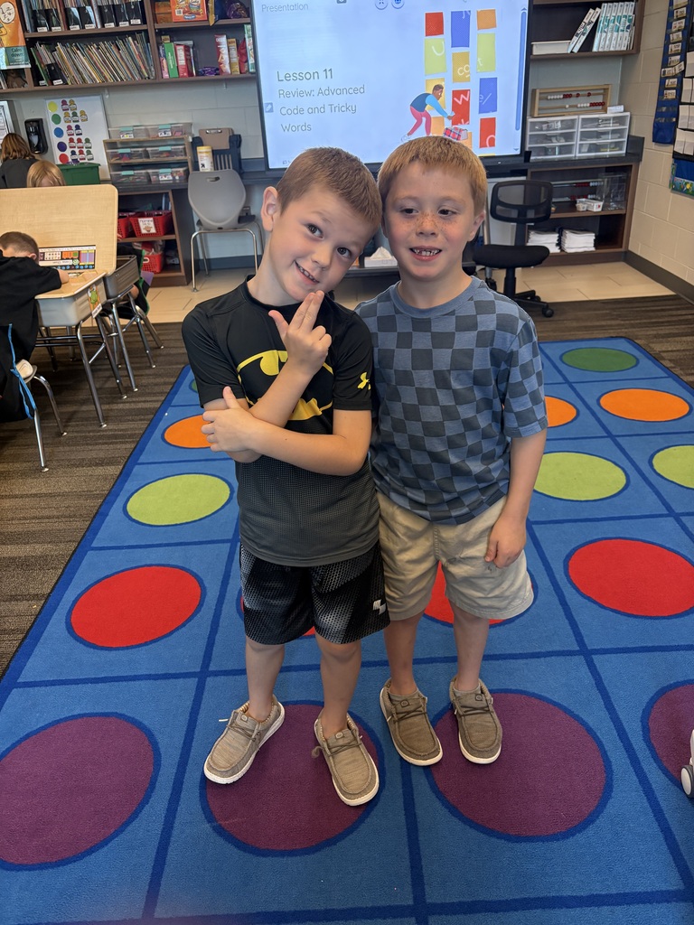 Two students stand together smiling on a colorful classroom rug during a lesson on advanced code and tricky words.