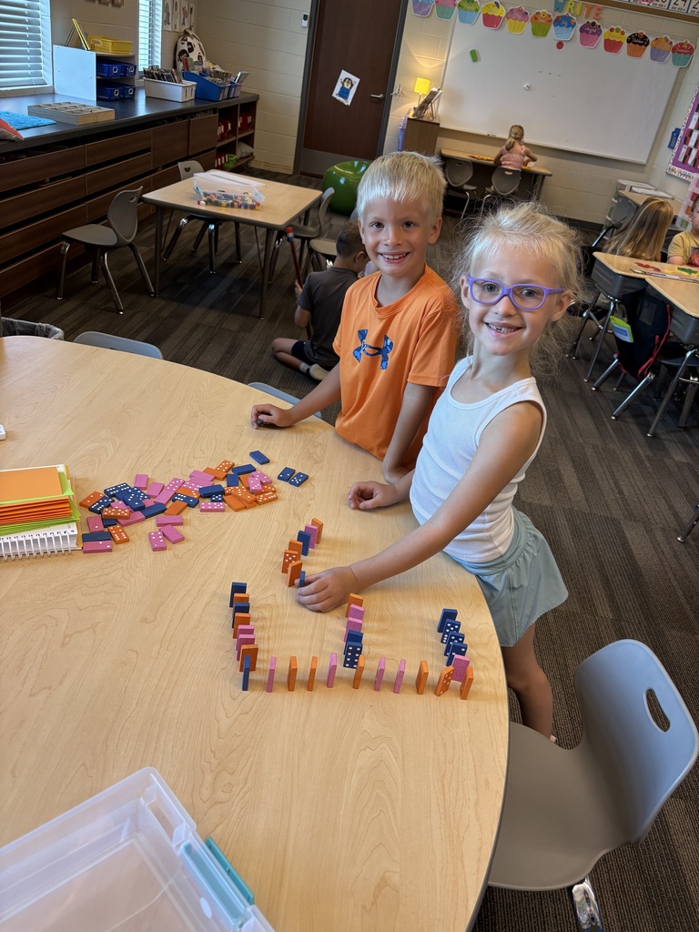 Two smiling students arrange colorful dominoes in lines and patterns across a classroom table.