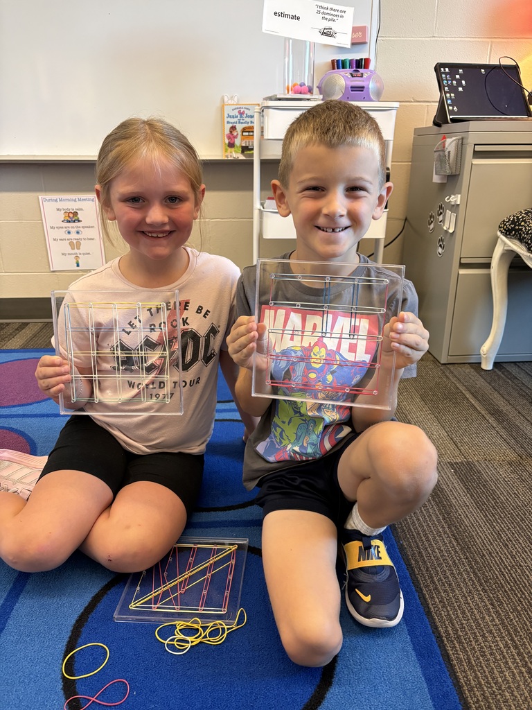 Two smiling students display clear geoboards with colorful rubber-band designs while seated on a classroom rug.
