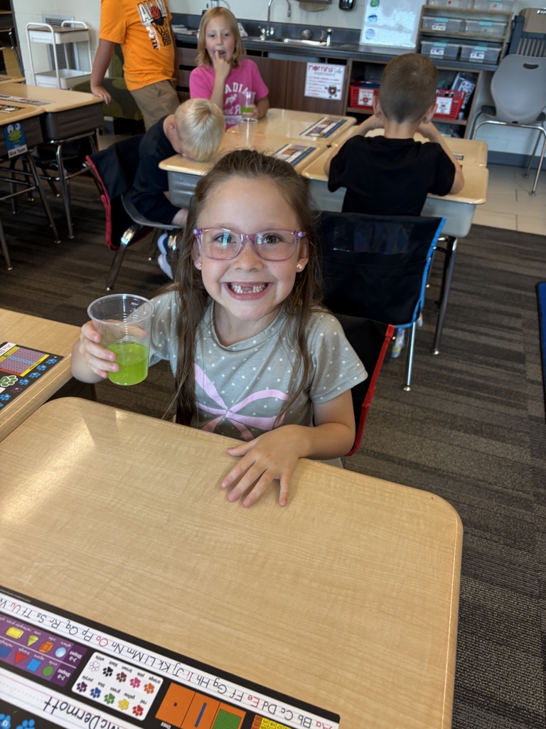 Smiling student with glasses holds a cup of bright green liquid while seated at a classroom desk.