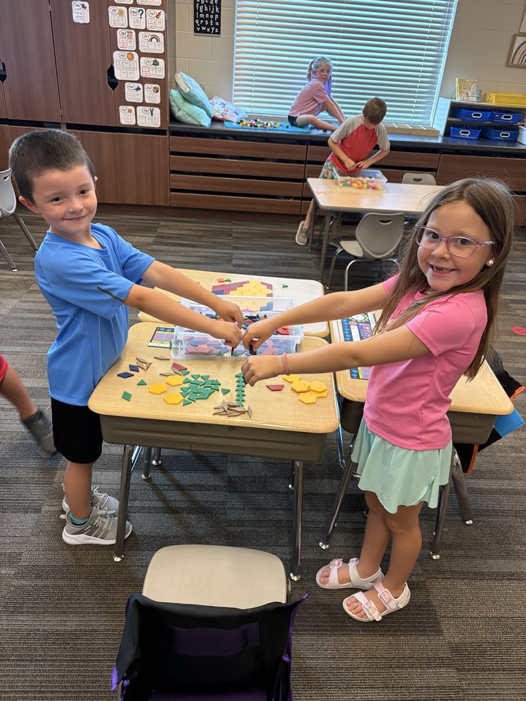 Two students smile while exploring colorful geometric pattern blocks spread across a classroom desk.