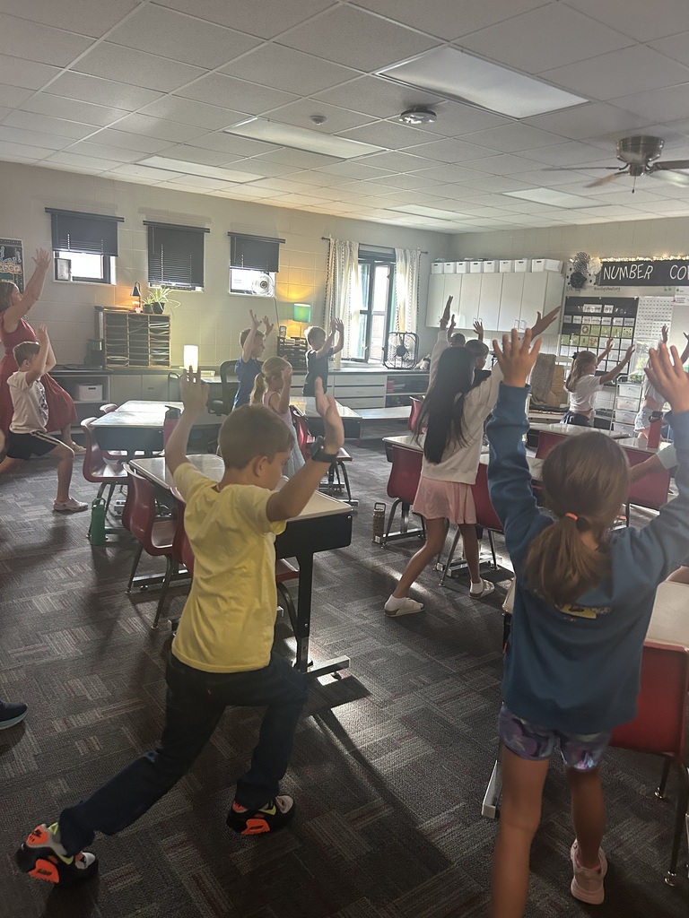 Students stand throughout the classroom with arms raised, participating together in a movement activity.