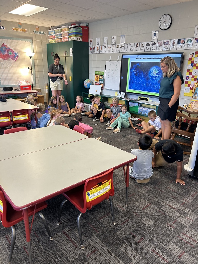 Students sit in a circle on the classroom floor with two teachers during a group lesson.