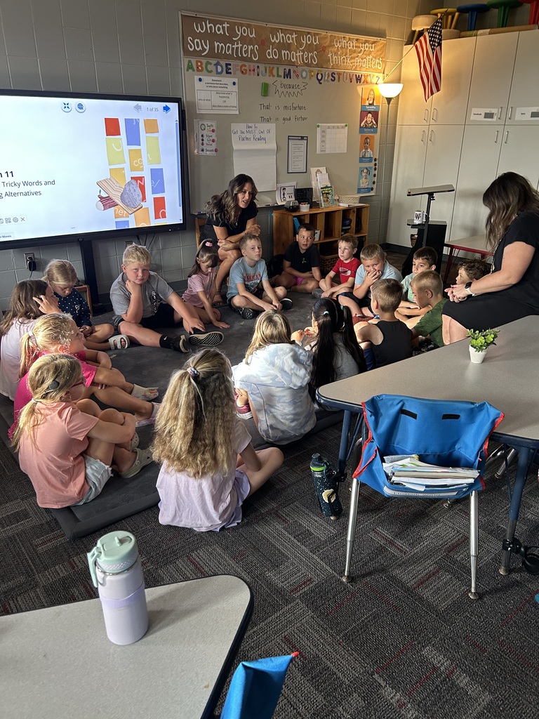 Students gather on a classroom rug with teachers for a group discussion near an interactive display.