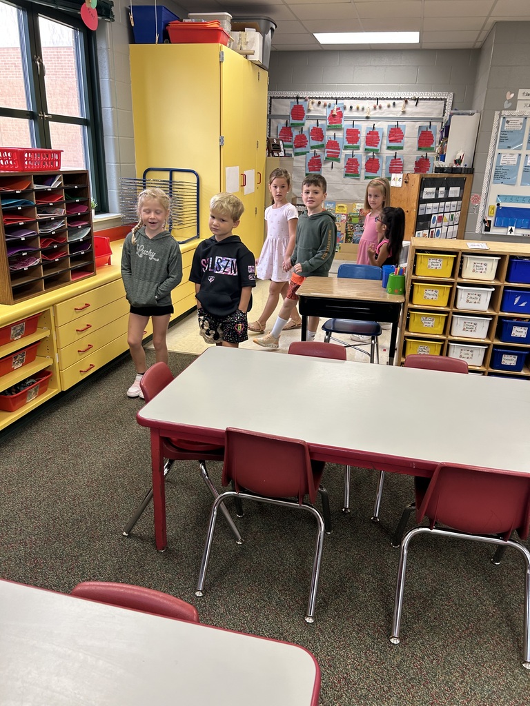 Students stand together near colorful classroom storage and tables during a group activity.