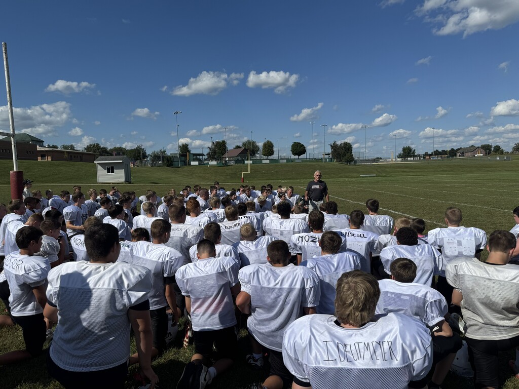 Football players in white practice jerseys kneel together on a sunny field while listening to a coach.