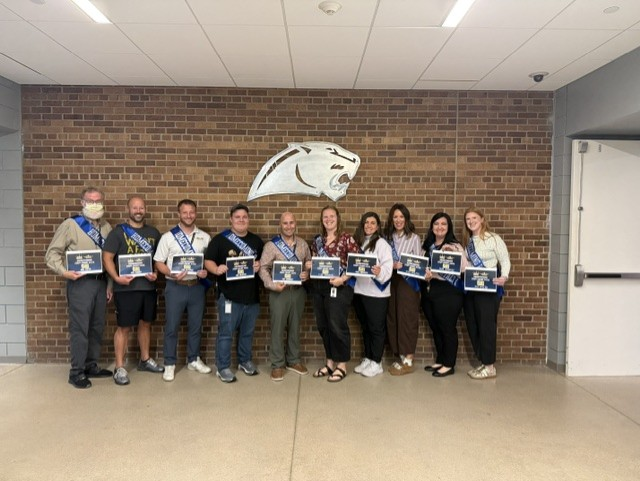 Ten school staff members stand beneath a cougar logo, smiling and holding recognition certificates.