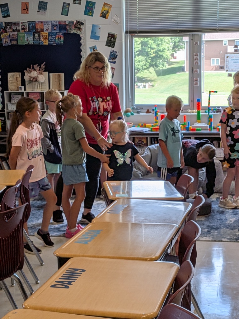 A teacher guides students through a classroom activity as children stand together among desks and learning materials.