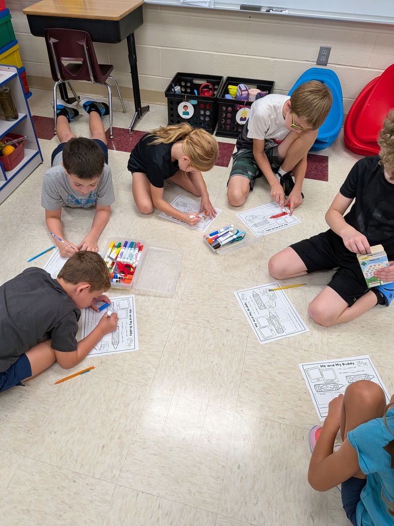 Students sit on the classroom floor completing “Me and My Buddy” worksheets with markers and pencils.