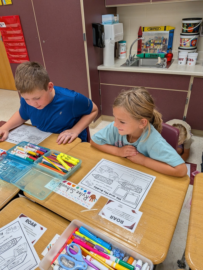 Two students work together at desks on a buddy activity sheet surrounded by colorful classroom supplies.