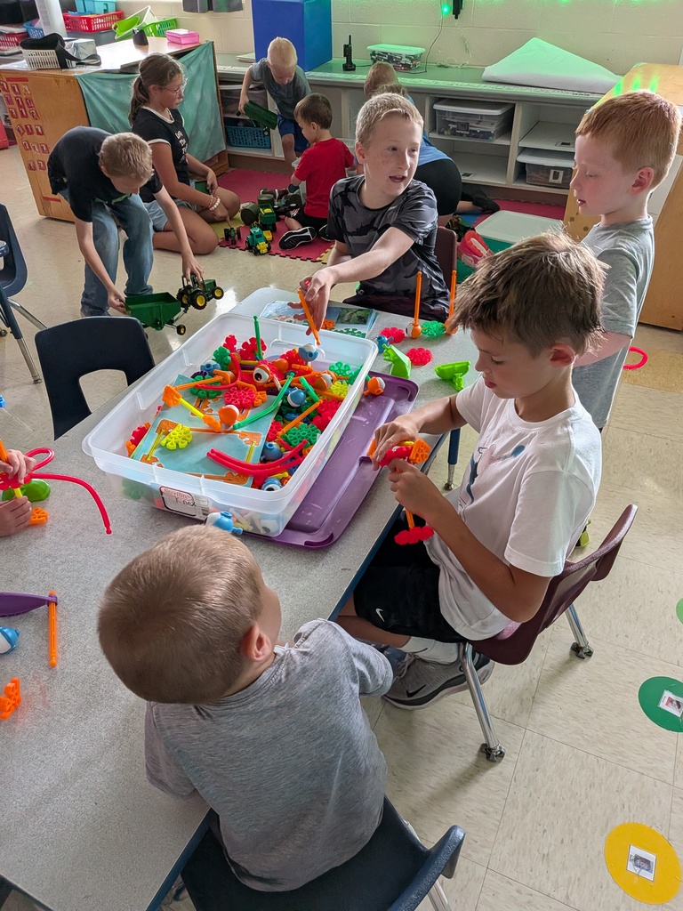 Students gather around a classroom table, building colorful creations while others play with toy tractors nearby.