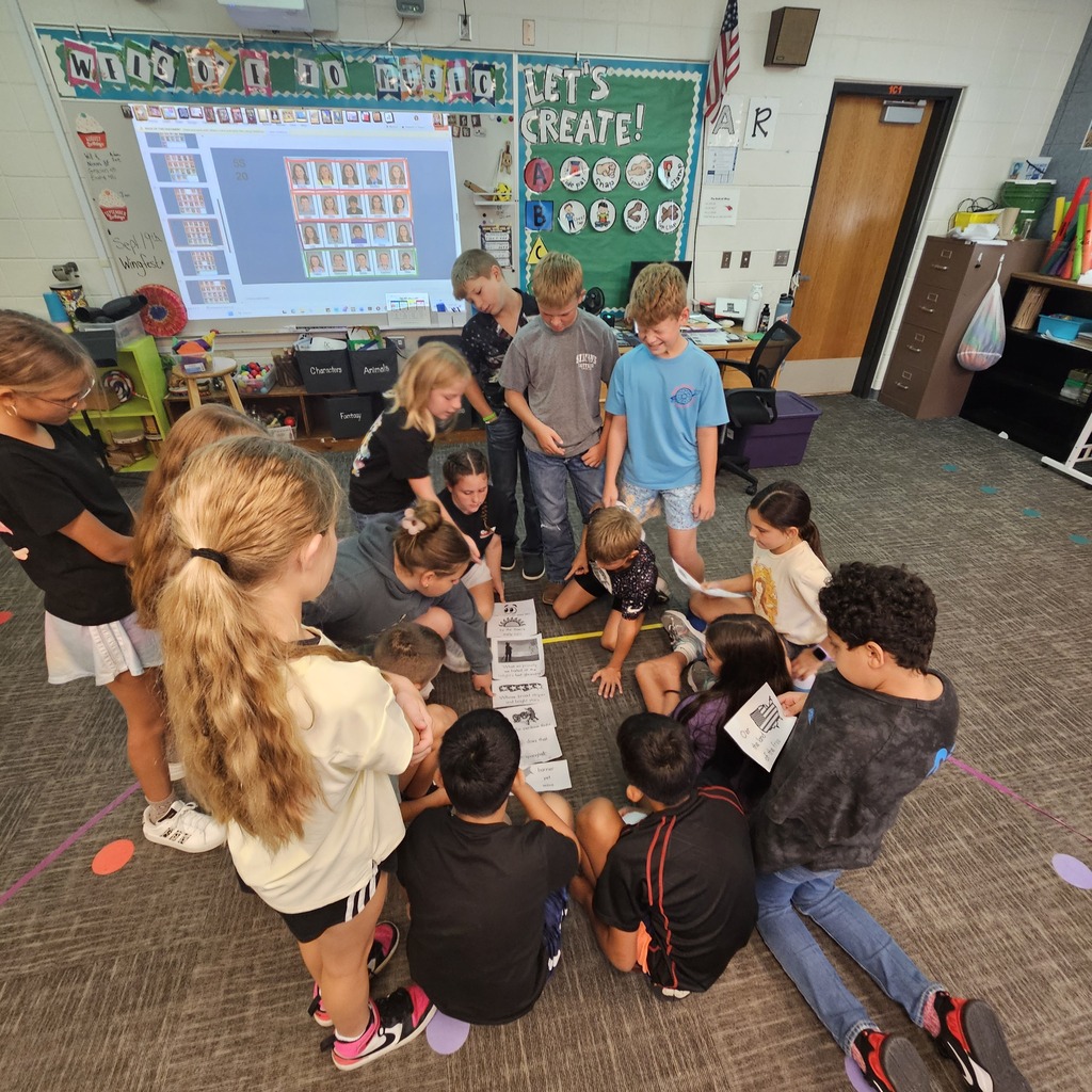 Students sit and stand together, collaborating on an illustrated sequencing activity spread across the classroom floor.