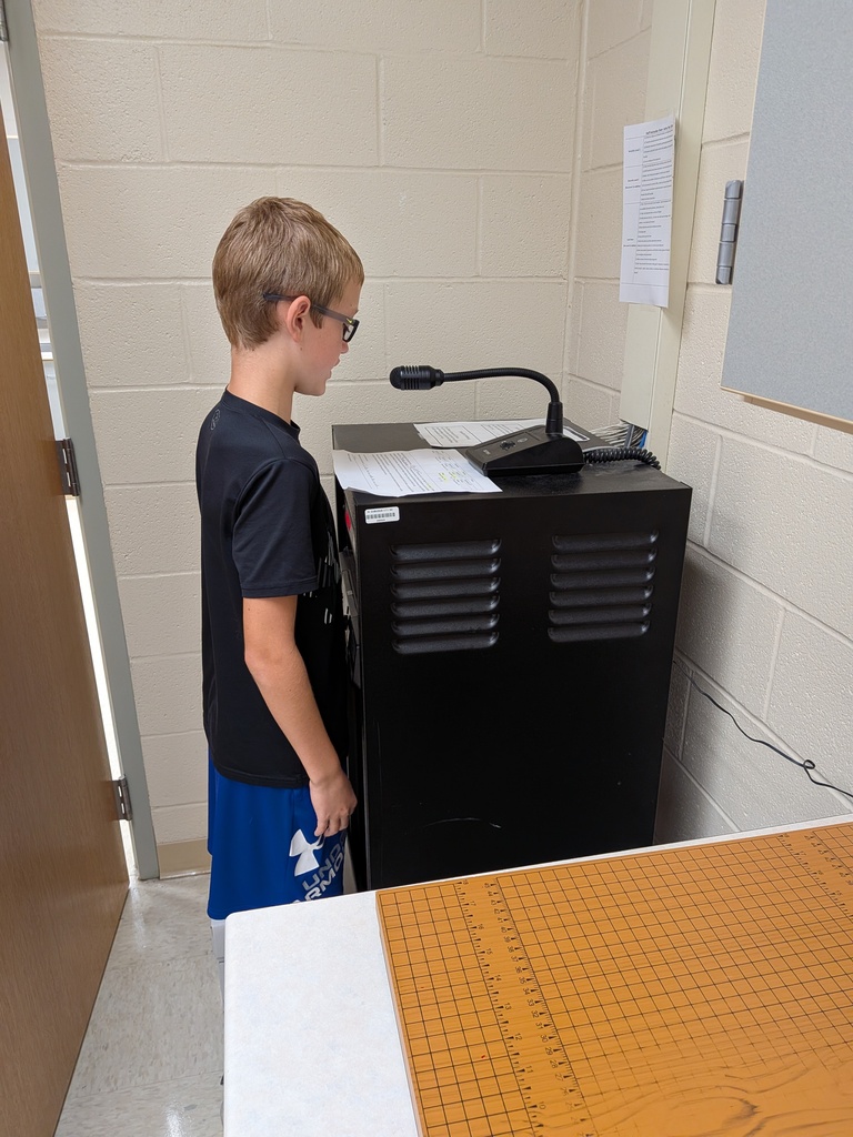 Student wearing glasses reads from a script while standing at a microphone connected to classroom equipment.