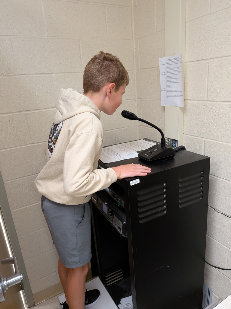 Student reads from a printed script while speaking into a microphone on top of classroom audio equipment.