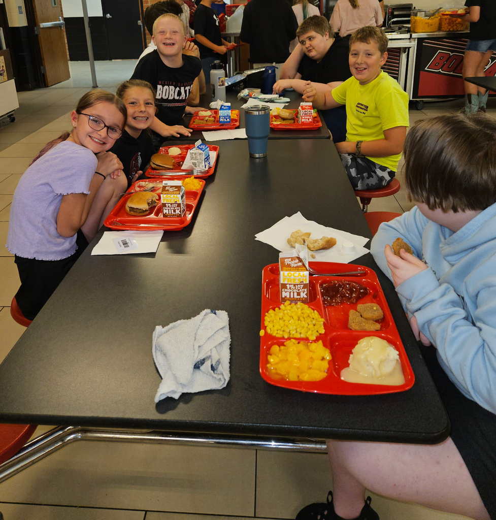 Students sit together at a cafeteria table, smiling and eating school lunches from red trays.