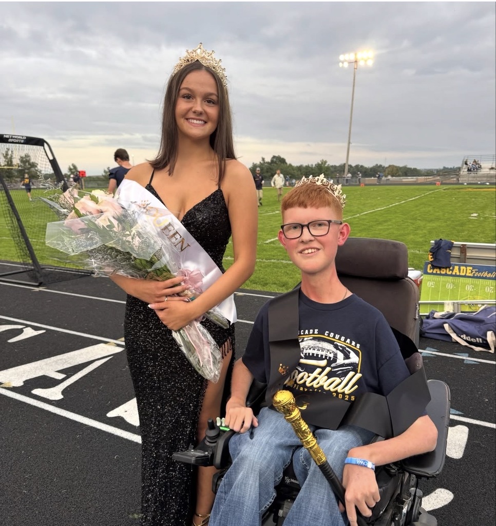 Two crowned students pose on a football field, one holding flowers and the other seated in a power wheelchair.