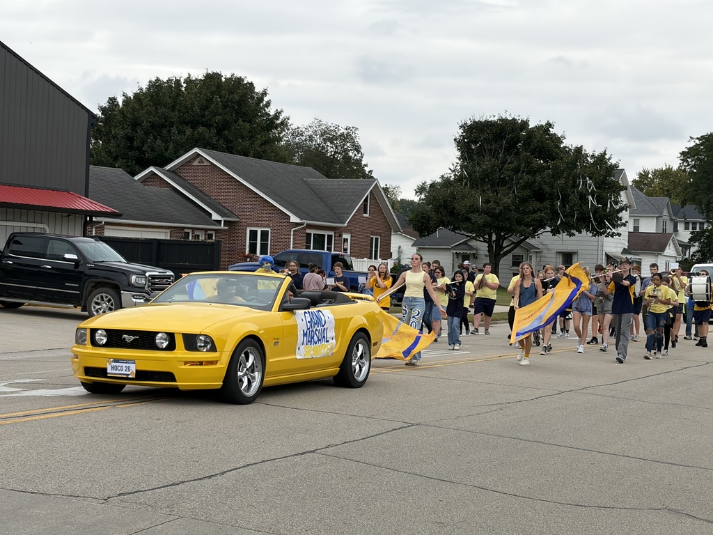 A yellow convertible leads students, color guard, and marching band members along a street during a school parade.
